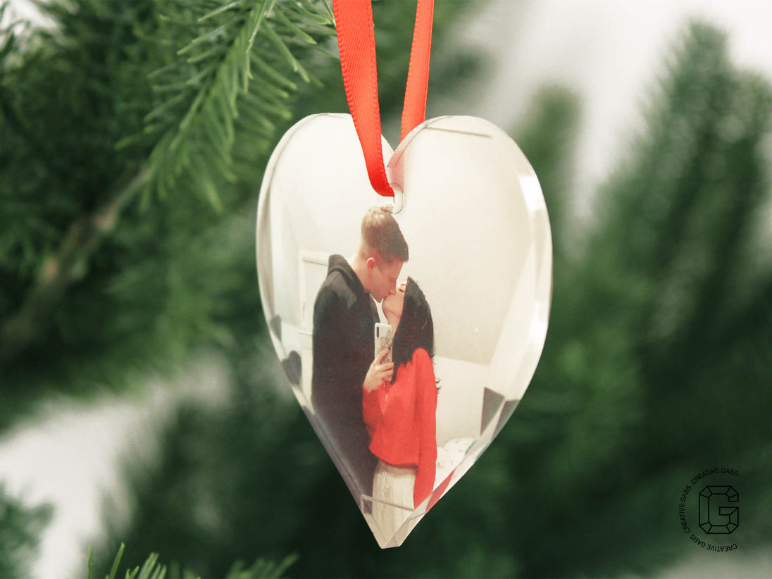 Nahaufnahme einer herzförmigen Acryl-Weihnachtskugel mit Foto – aufgehängt am Tannenbaum mit rotem Band