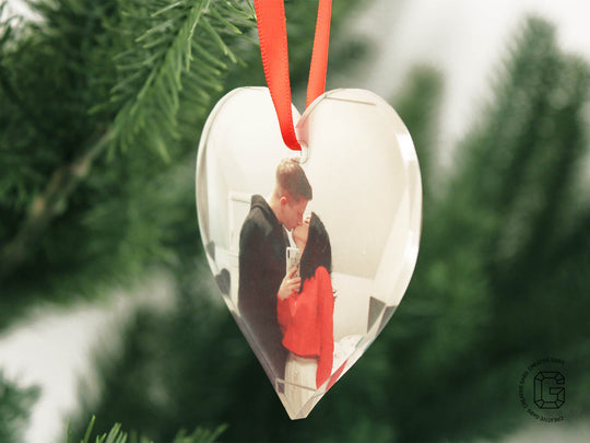 Nahaufnahme einer herzförmigen Acryl-Weihnachtskugel mit Foto – aufgehängt am Tannenbaum mit rotem Band