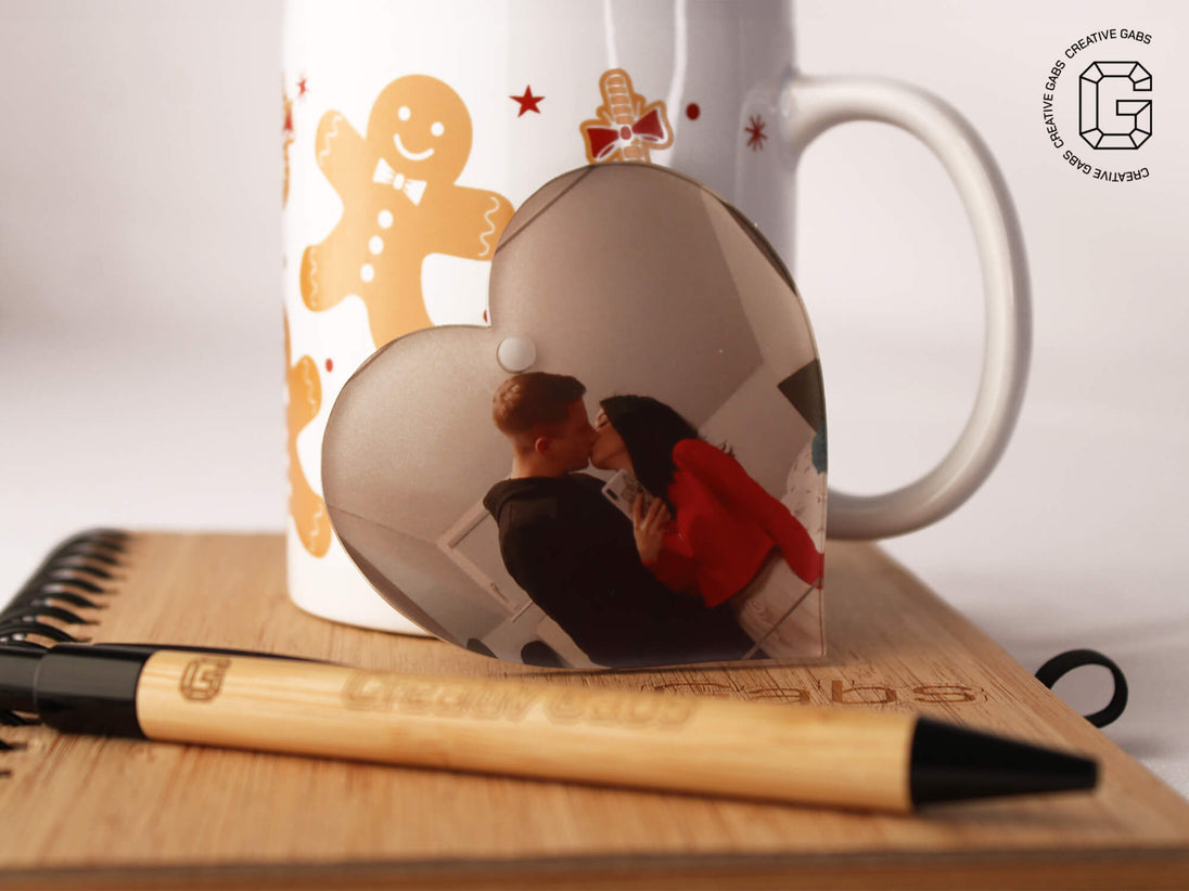 Herzförmige Acryl-Weihnachtskugel mit Foto – Größenvergleich neben weihnachtlicher Tasse und Kugelschreiber