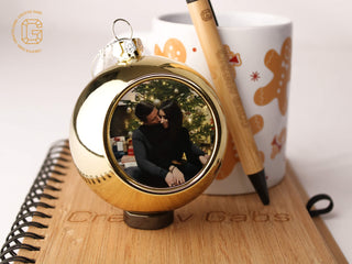 Personalisierte goldene Weihnachtskugel mit Foto im Größenvergleich neben Tasse und Kugelschreiber